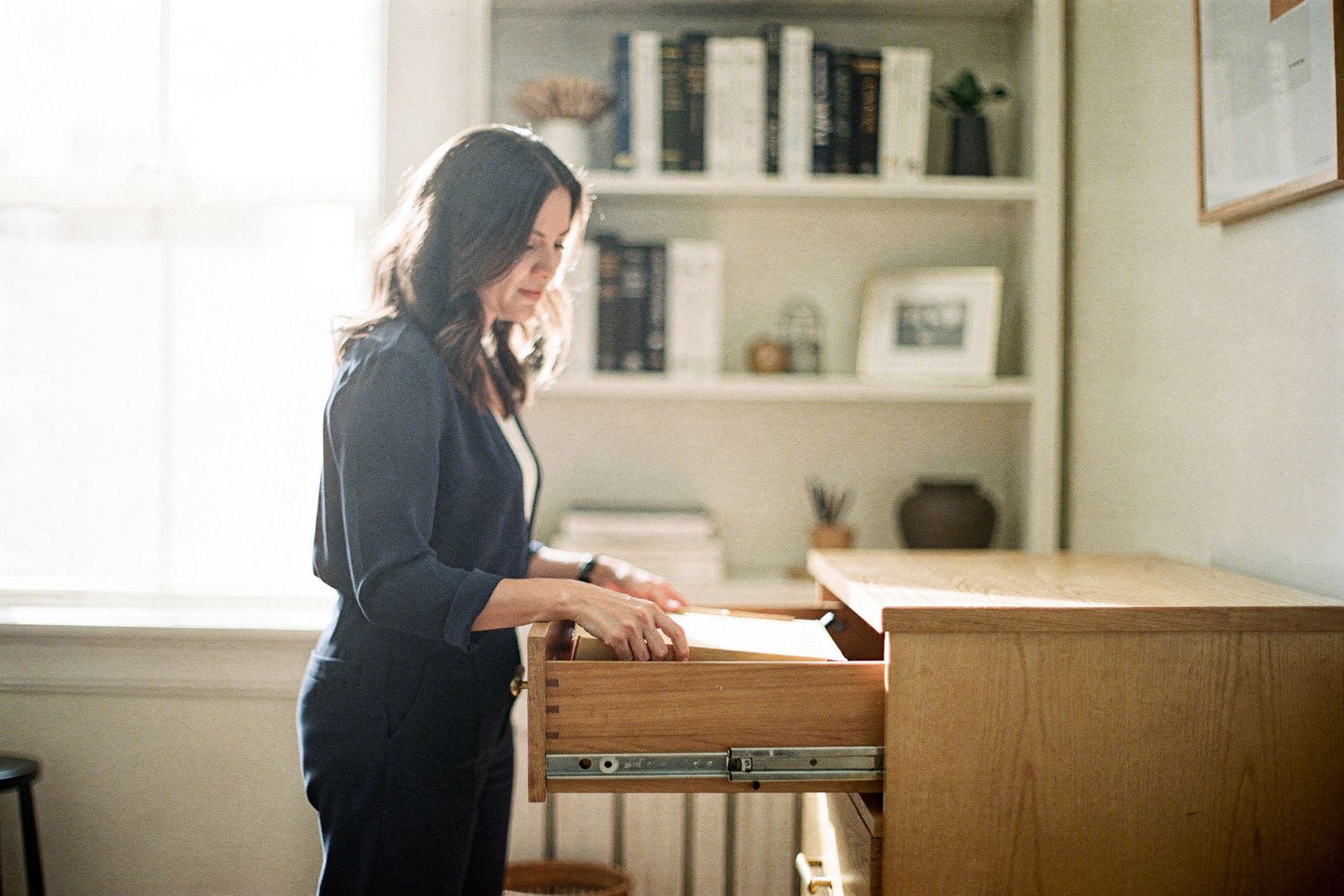 A woman in her thirties filing a paper folder into a wooden filing cabinet in a calm home office, bookshelves behind her.
